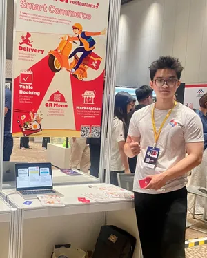 Đức in a white Savyu T-shirt and an exhibitor lanyard at the Savyu booth, beside a roll-up banner and a laptop.