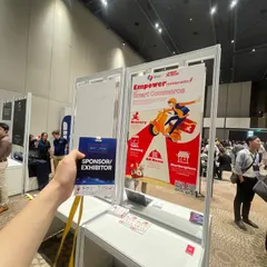 A hand holding a French Tech Summit Vietnam sponsor and exhibitor badge in front of the Savyu roll-up banner.