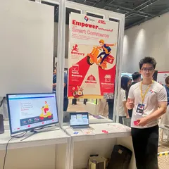 Đức in a white Savyu T-shirt and an exhibitor lanyard at the Savyu booth, beside a roll-up banner and a monitor showing Giaodoan.vn.