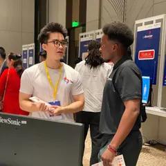 Đức, holding flyers, talks with a visitor in a dark polo shirt among the exhibition booths.